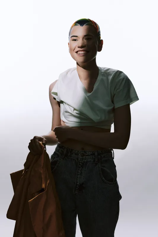 Young woman with short hair smiling, dressed in casual white shirt and jeans, holding a brown jacket, standing against a bright neutral background.