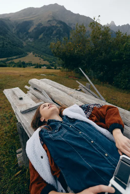Woman lying on a wooden bench in a mountain meadow, relaxed and contemplative in a denim jacket holding a camera. Scenic landscape and peaceful outdoor retreat with distant peaks.