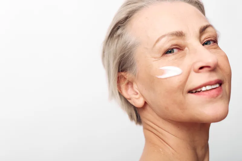 Smiling mature woman with short blonde hair applying white face cream on cheek. Close-up on light background. Concept of skincare, antiaging, wellness, and natural beauty care.