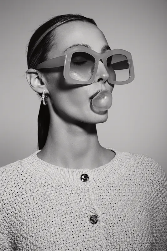 Portrait of a white woman with fair skin wearing large sunglasses and blowing bubble gum. Fashion concept with stylish accessories in monochrome studio.