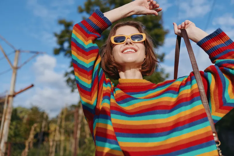 woman rainbow sweater sunglasses smile outdoors fashion portrait of young adult with shoulder bag, colorful casual style on a sunny day, joyful and carefree mood