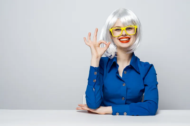 Эмоциональная женщина с косметикой, красными губами и белым париком делает жест руками, на ней синяя рубашка