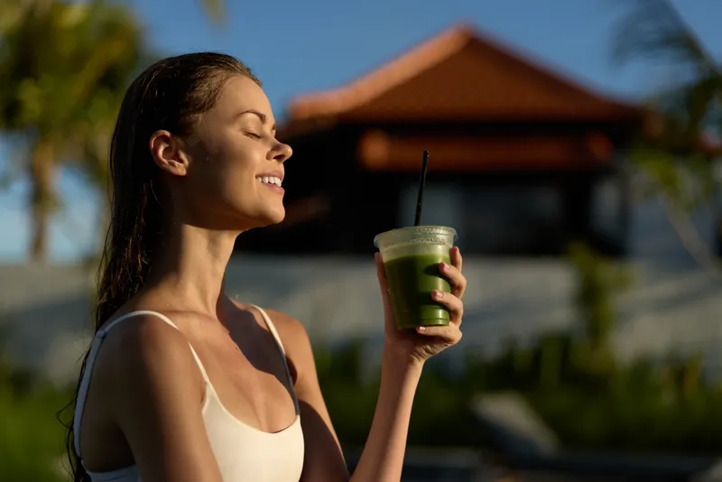 Woman outdoors enjoying green smoothie, smiling with eyes closed, sunlight on face, relaxed mood, healthy lifestyle, natural background with blurred architecture and palm trees