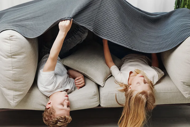 Two children lie under a gray blanket on a beige sofa, playing and smiling in a cozy home environment. Concept of childhood fun, comfort, and family bonding moments.