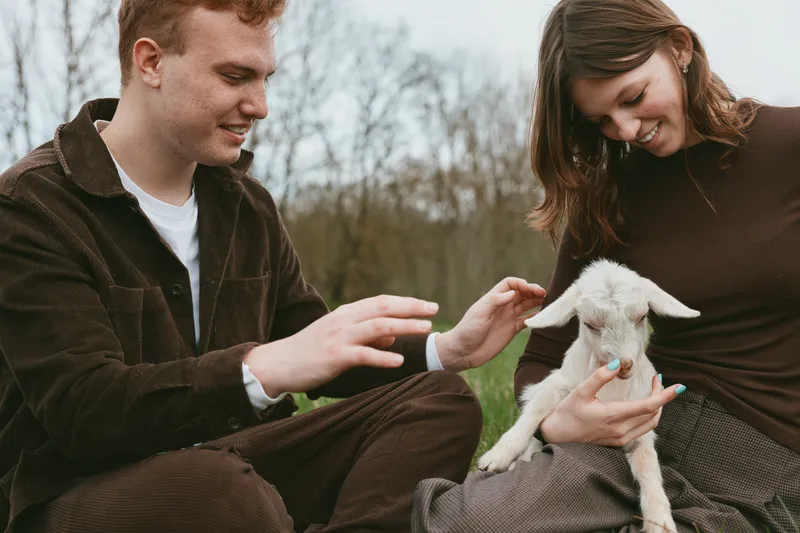 Two adults share a gentle moment outdoors with a curious goat kid, smiling and petting the animal in a casual lifestyle scene, daytime, natural park setting