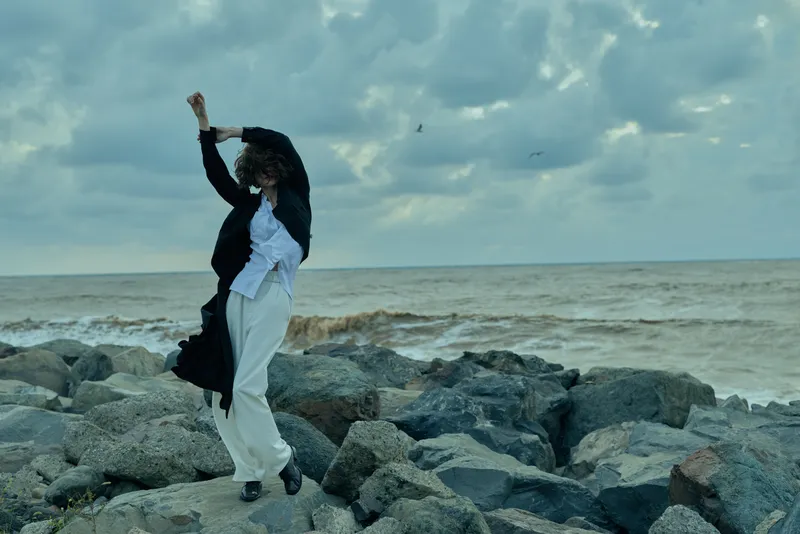 Woman dances on the rocks along the sea, coat fluttering, wind sweeping, outdoor scene with overcast sky, conveying movement, balance and style in a rugged coastal setting
