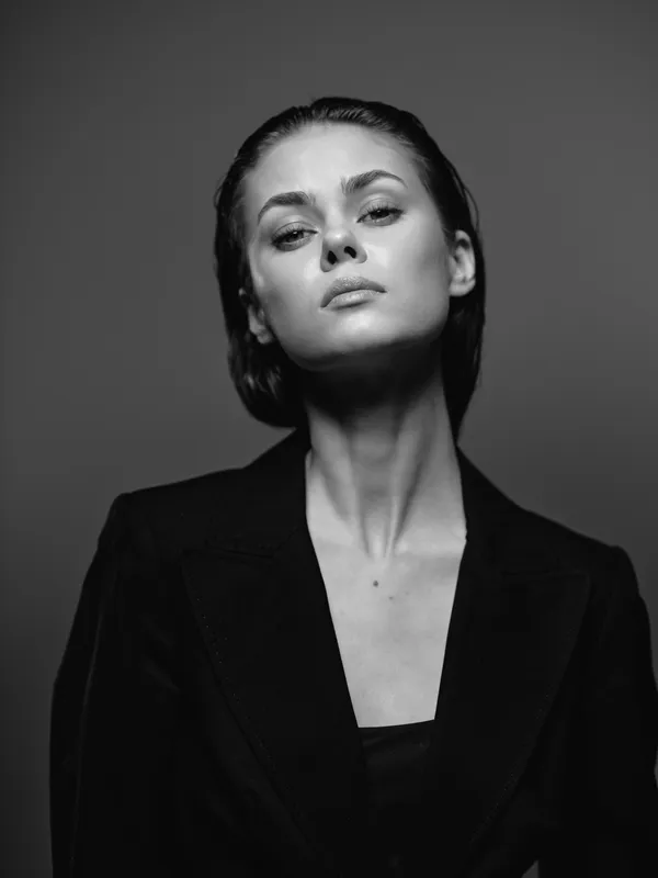 Black and white portrait of a confident woman in dark clothing with serious expression and dramatic lighting.