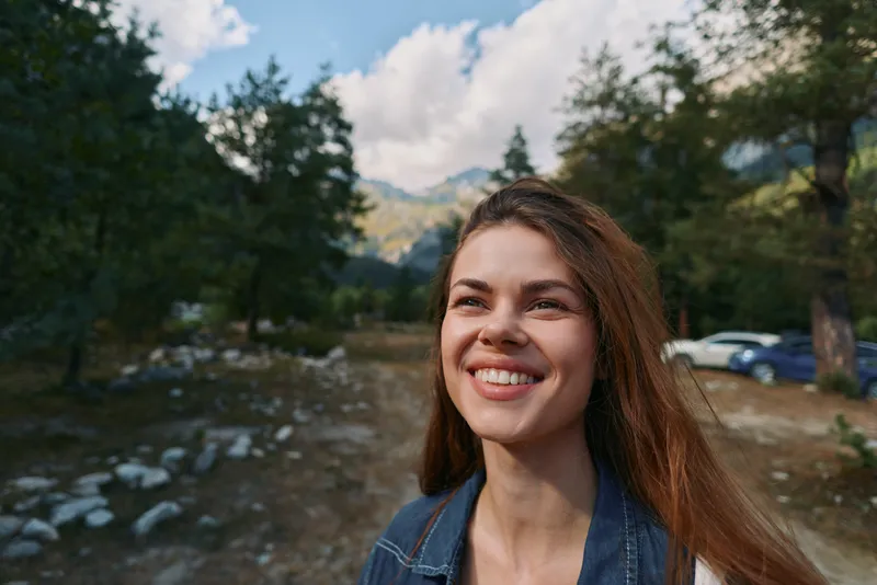 A cheerful woman stands outdoors in a natural setting, smiling brightly in sunlit weather, wearing casual denim and a light jacket, creating a warm, inviting portrait of everyday happiness.