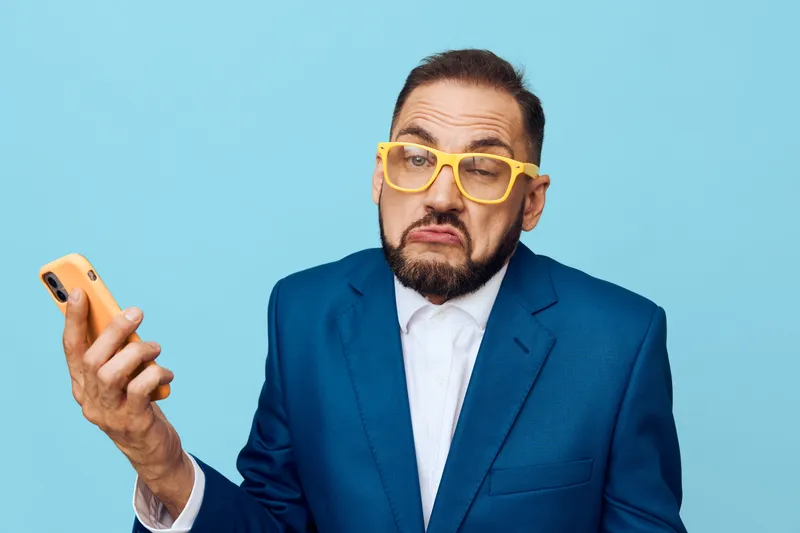 A professional man in a blue suit stands against a solid colored background, expressing doubt and concentration as he checks his phone.