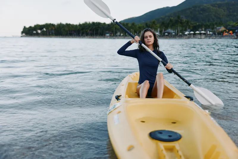 A young woman paddles a yellow kayak across calm water, her focused posture and steady stroke conveying momentum and adventure in a serene lakeside setting.