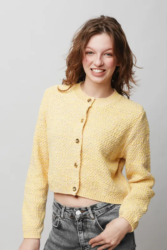 Smiling young woman with curly hair wearing a yellow knitted cardigan and grey jeans stands against a light background. Casual fashion, happiness, and natural beauty concept.