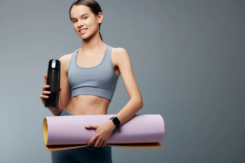 Female fitness enthusiast in athletic wear holding a water bottle and yoga mat, ready for a workout, showcasing balance, strength, and an active lifestyle in a studio.