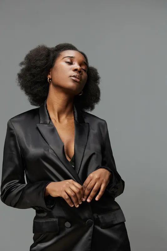 Confident young Black woman in a stylish black satin blazer, posing against a neutral gray background, exuding elegance and sophistication