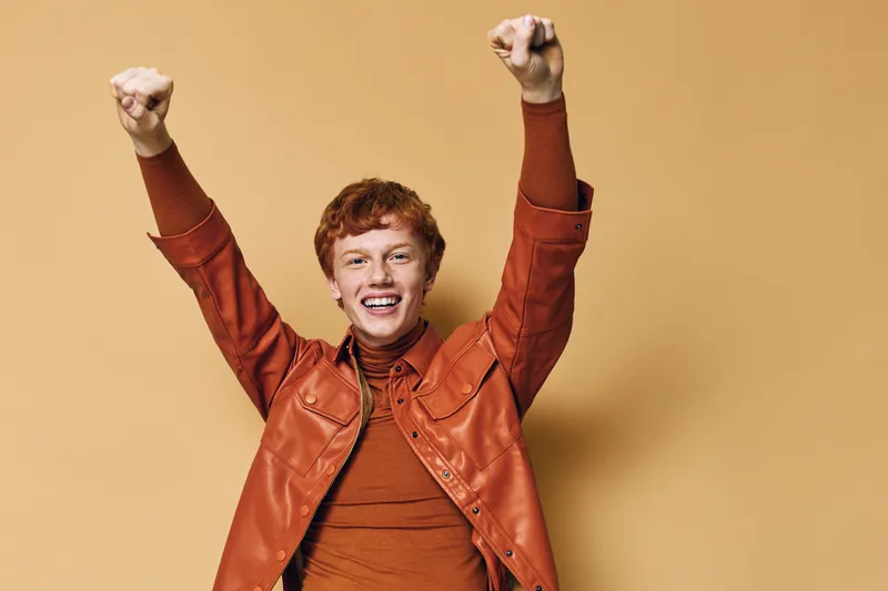 Young man with red hair wearing orange jacket and sweater raising hands with excitement and smile on beige background in studio. Celebration, happiness and joy with casual style clothing.
