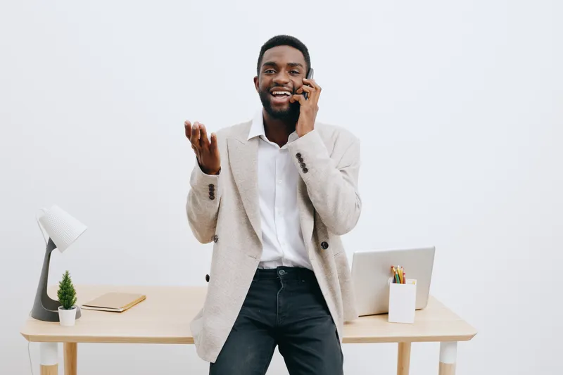 Smiling cheerful man talking on mobile phone in bright office, casual business attire, standing near desk with laptop and stationery, professional work environment, positive mood. People lifestyle con