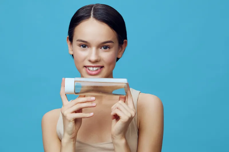 Smiling young woman holding a clear skincare bottle against a blue background, highlighting beauty and self care concepts with a fresh and vibrant aesthetic