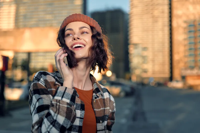 woman phone smile city beanie plaid sunlight portrait captures candid lifestyle and authenticity during golden hour glow; mindful living moment of emotional storytelling as she chats outdoors