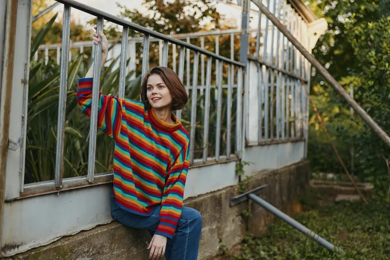 Woman in colorful striped sweater sitting by a greenhouse, portrait of smiling young adult with plants, casual fashion, outdoor lifestyle and relaxed autumn mood.