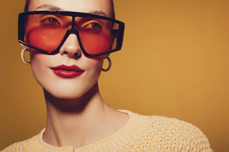 Stylish woman with oversized orange sunglasses and gold hoop earrings poses against a warm yellow background. Fashion portrait with bold accessories and confident expression.