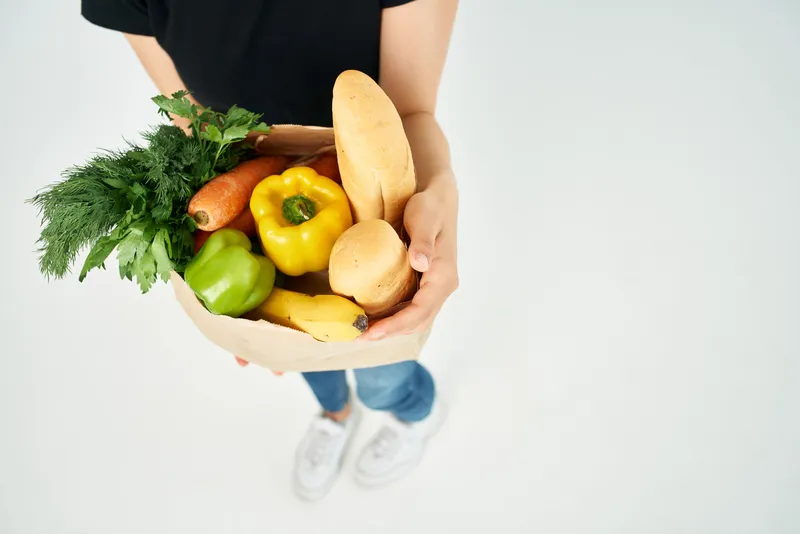 Крупным планом показана женщина, держащая в руках пакет с продуктами, наполненный овощами, что ассоциируется со здоровым питанием