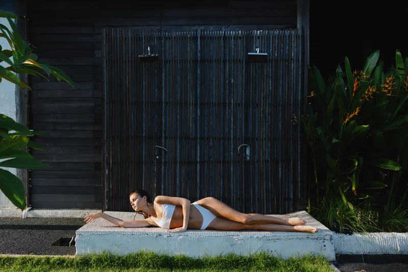A young woman in a white swimsuit lounges on a concrete ledge beside a dark wooden wall and a waterfall, creating a calm, sunlit lifestyle moment outdoors.