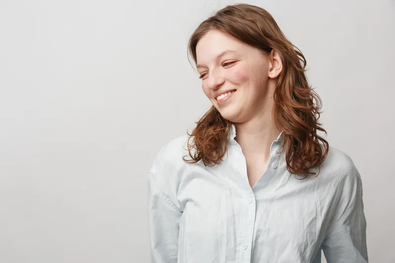 Smiling young woman with curly brown hair wearing white shirt looks down shyly on light background. Concept of natural beauty, happiness, casual style, and positive emotions.