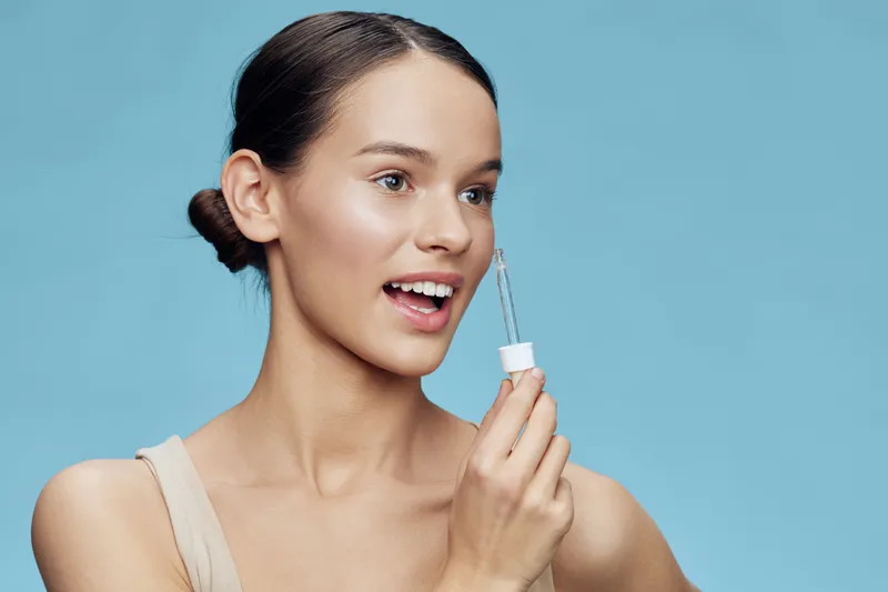 Young woman applying skincare serum on a bright blue background, showcasing a healthy skincare routine with a joyful expression Natural beauty concept