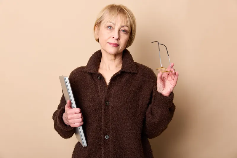 Elderly woman grooming with a hairbrush, holding a notebook, standing in a beige studio background, home selfcare concept. Soft warm light, calm mood.