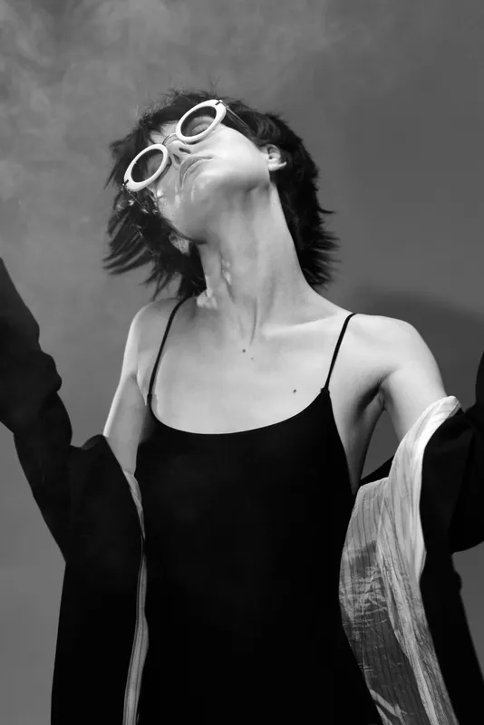 Black and white photo of a young woman wearing sunglasses and a dress, posing with closed eyes and relaxed expression.