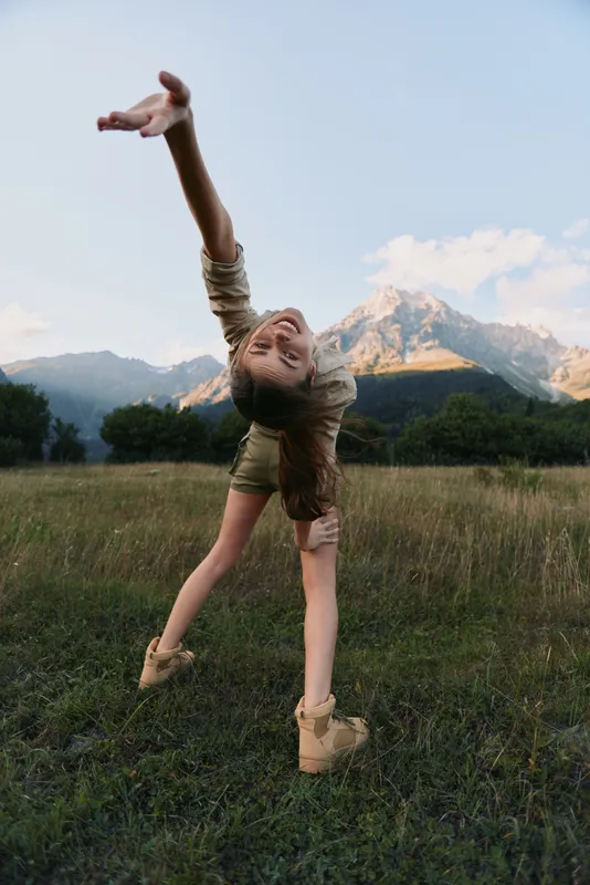 Young girl dances in a sunlit meadow with mountains in the background, arms raised in joy