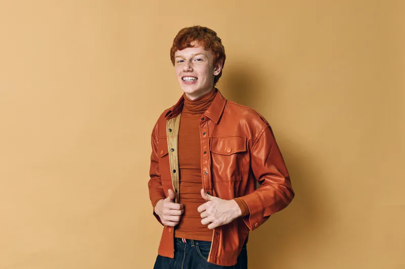 Young man with red hair wearing brown leather jacket and turtleneck sweater standing against beige background smiling at camera with casual style and confident pose.
