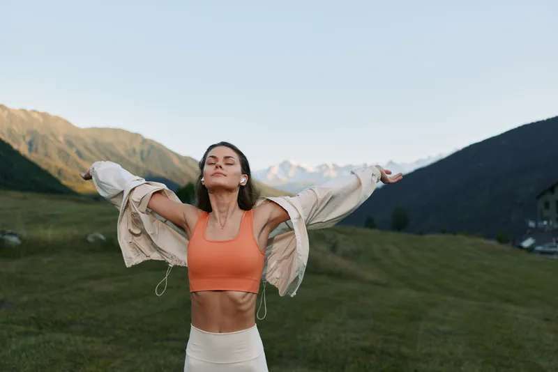 Joyful woman stands outdoors with arms wide, wearing an orange athletic top and light jacket. Sunlit mountains form a scenic backdrop, conveying energy, freedom, and vibrant lifestyle.