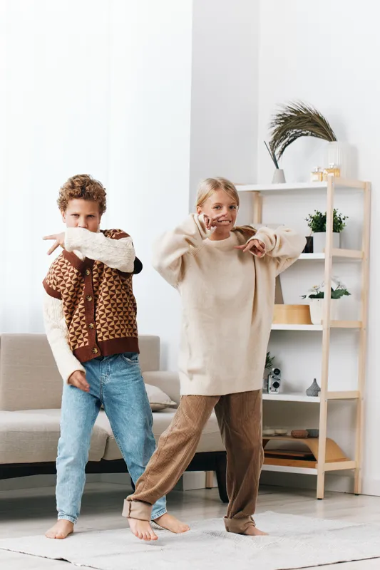 Authentic lifestyle image of two children indoors, dressed casually, engaged in playful movement that captures daily learning through shared activity and genuine joy.