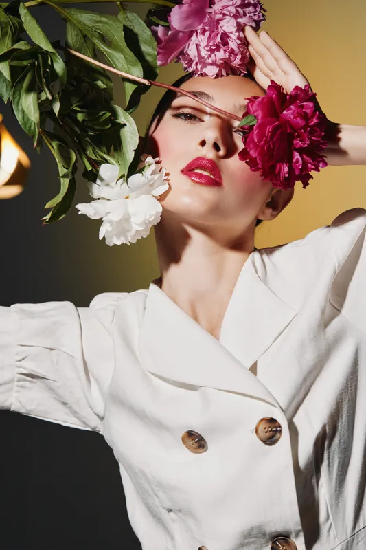 Portrait of a stylish woman dressed in a white jacket holding pink and white flowers near her face, bright makeup with red lipstick, creative beauty and fashion concept.