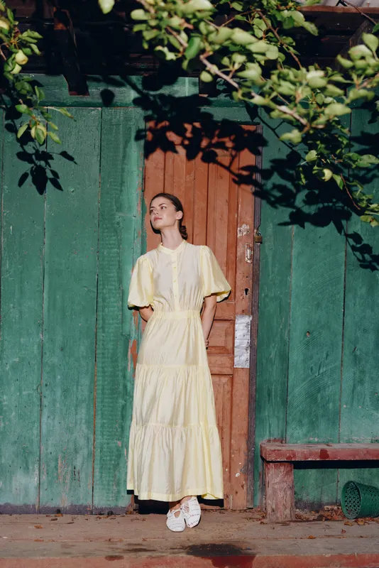 Portrait of a woman in a pale yellow dress standing beside a weathered green door in a sunny outdoor scene, conveying calm elegance and vintage charm.