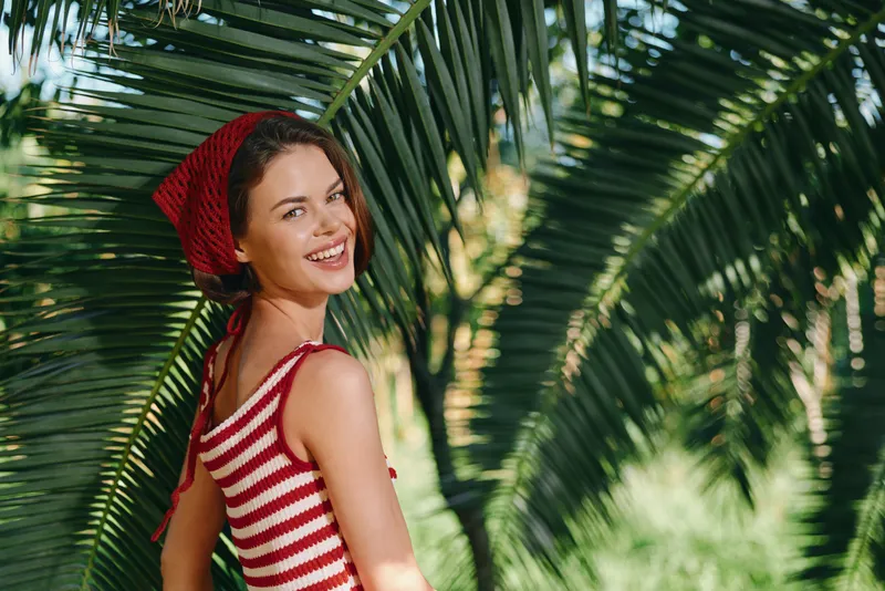 A cheerful woman stands among lush palm leaves, wearing a striped red and white top with a headscarf. She smiles brightly, exuding relaxed vacation vibes and warm sunshine.