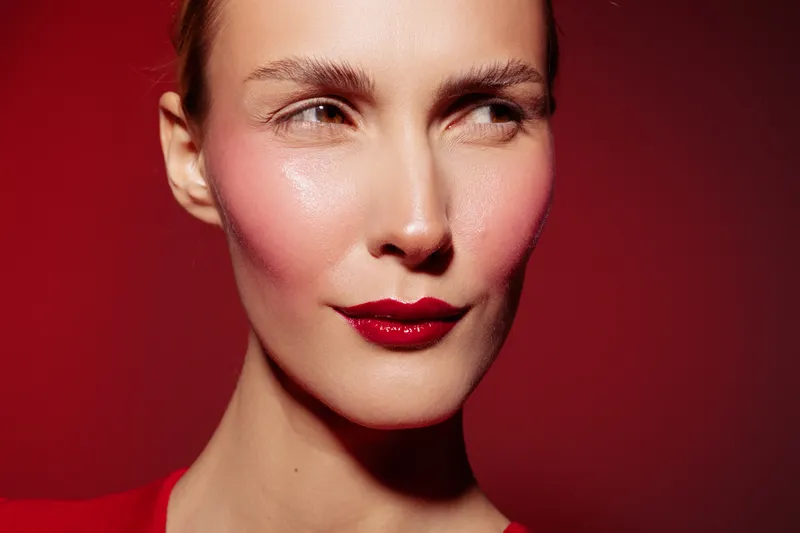 Close-up of a woman with red lipstick and pink blush on her cheeks looking sideways. The red background and lighting create a bold and dramatic beauty portrait concept.
