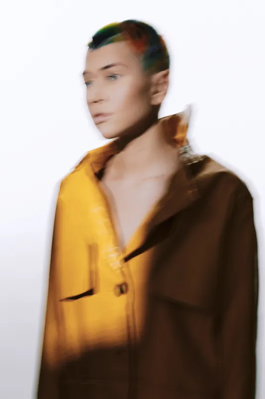 Blurred woman with short hair wearing a yellow jacket, standing against a white background, featuring soft lighting and an artistic fashion style.