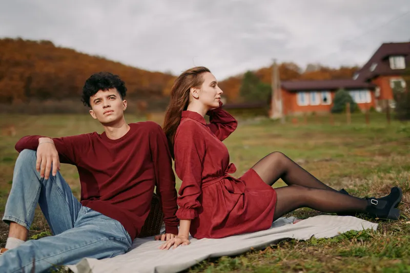 A young couple sits back to back on a blanket in an autumn field, a candid, natural portrait capturing quiet emotion, authenticity and understated credibility in a real moment.
