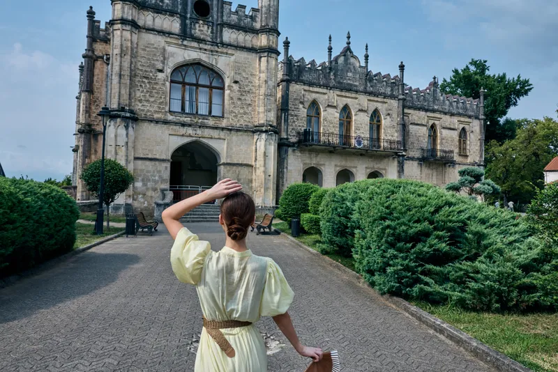 A woman in a pale dress stands on a gravel path facing a historic castle with manicured hedges, capturing a moment of travel and discovery.