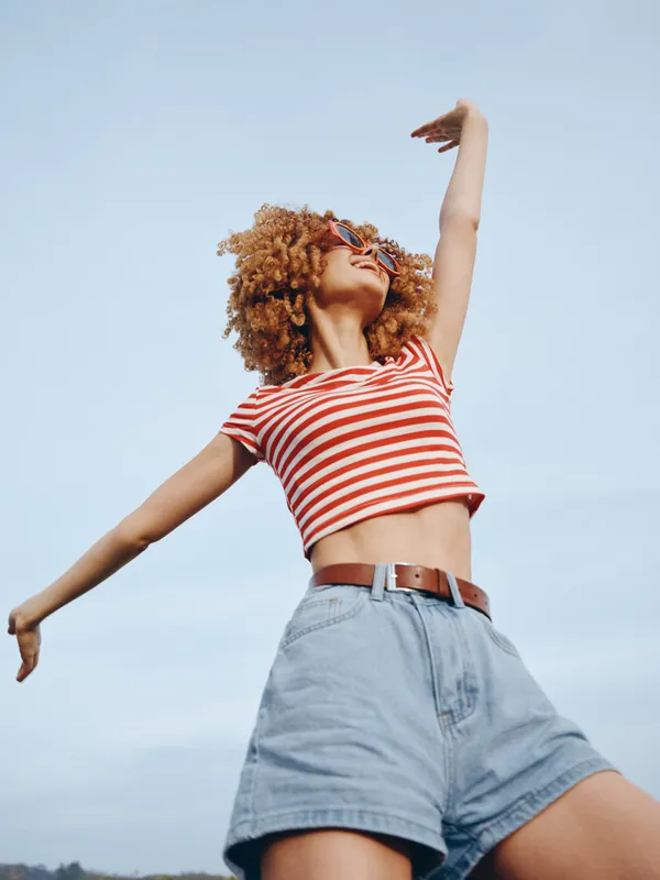 A young person wears a red striped crop top and high rise denim shorts, arms raised toward the sky, sunlit blue backdrop, creating a carefree summer fashion moment.
