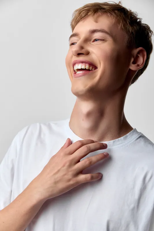 Smile, laughter and bright mood as a young man in a white shirt touches his chest, conveying joy, vitality and positive energy in a casual studio shot