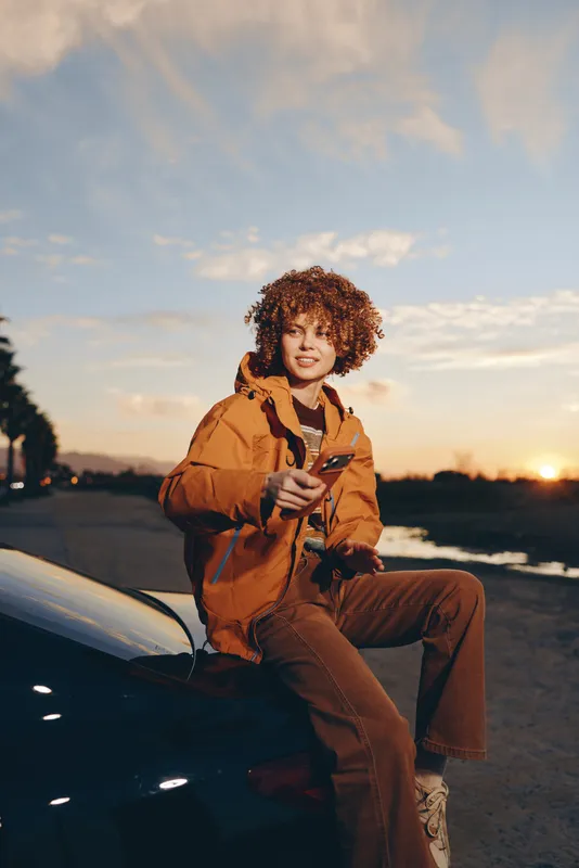 Woman lifestyle at sunset near a car, curly hair, casual jacket, holding a phone while sitting on the roadside, warm evening light, relaxed travel portrait, outdoor mood for women life