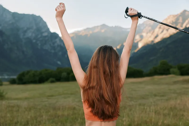 A woman stands in an open field with arms raised, embracing freedom and achievement against a scenic mountain landscape, conveying motivation and outdoor vitality.
