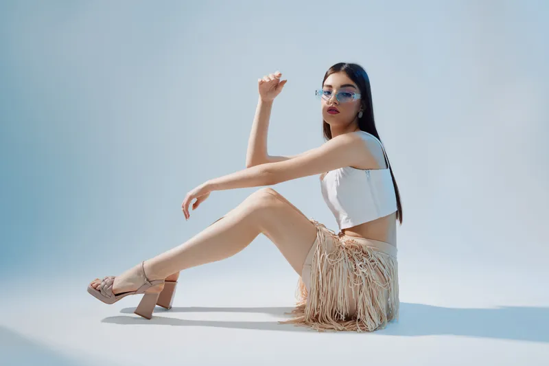 Young woman in trendy fashion outfit poses sitting on the floor against a light background. She wears stylish glasses and a fringed skirt. Concept of beauty, style, and fashion.