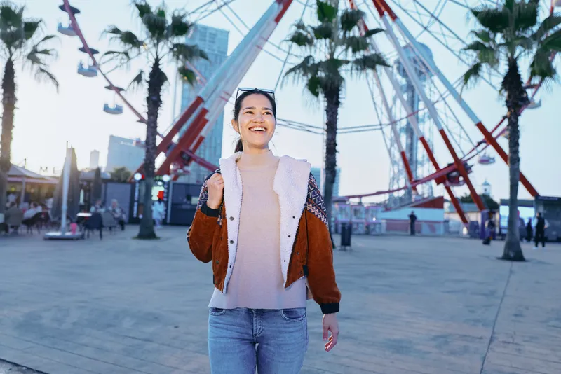 Woman smiles while walking in an outdoor plaza with palm trees and a modern sculpture, casual jacket and jeans, capturing urban leisure, travel vibe, and sunny everyday life