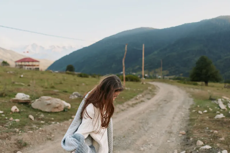 Young woman in a cozy knit cardigan walks along a rural dirt road with mountains in the background, capturing a calm, reflective lifestyle moment outdoors.