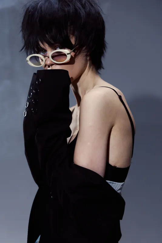 Stylish woman wearing sunglasses and black jacket posing against a dark background with a moody atmosphere.