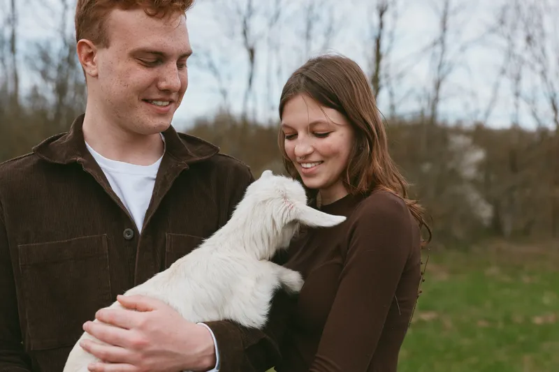 A couple gently holds a white goat kid in a sunlit meadow, smiling softly. The scene conveys pastoral lifestyle, animal bonding, and gentle affection in a natural outdoor setting.
