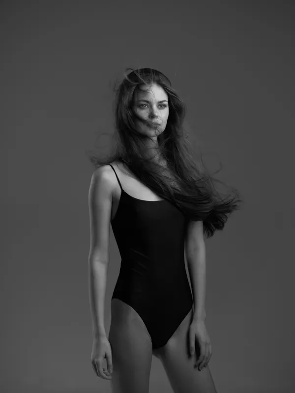 Black and white portrait of a woman in a black swimsuit with flowing hair and neutral expression against a plain background.